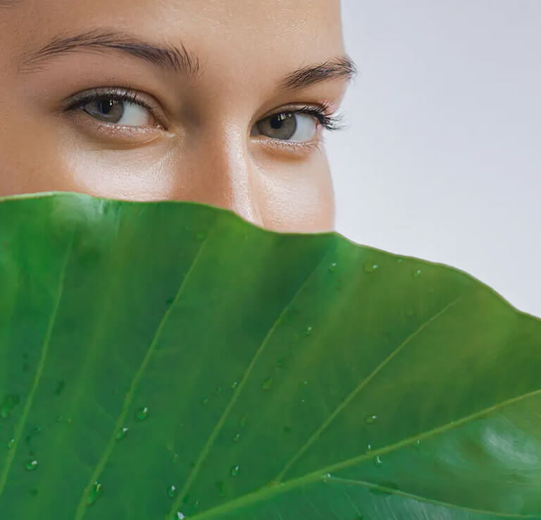 A woman with smooth skin peaks out from behind a leaf.