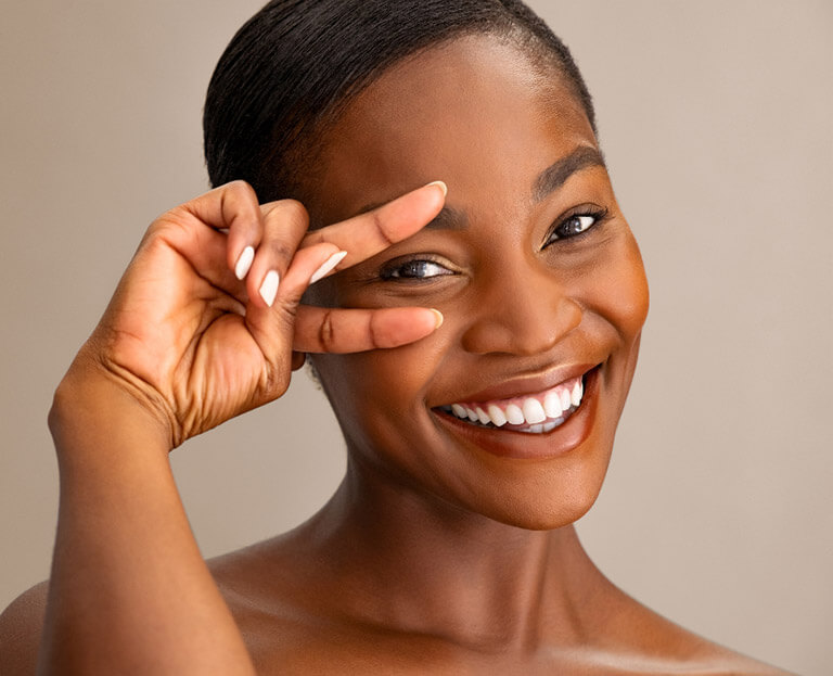 A black woman with flawless skin smiles at the camera. She is holding two fingers near her eye.