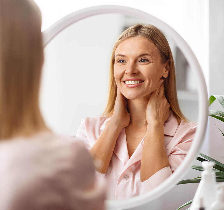 Cheerful middle aged woman with clear skin looks in mirror.